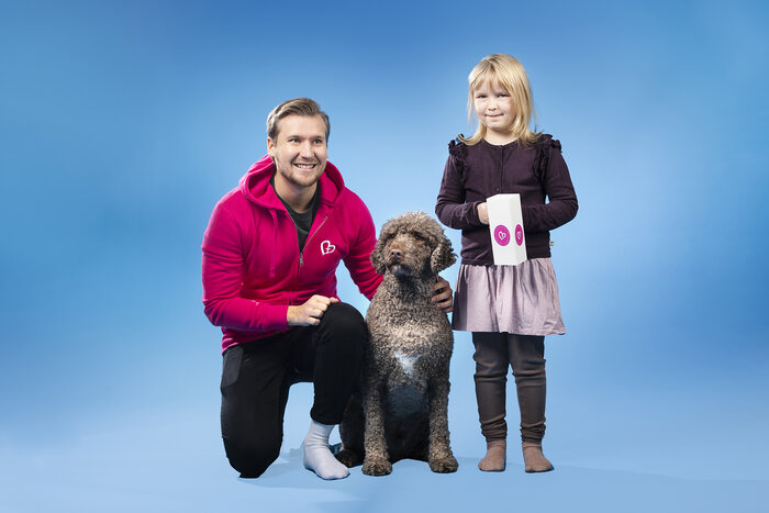 Hymyilevä mies silittää istuvaa koiraa, vieressä tyttö jolla kädessä keräyslipas.